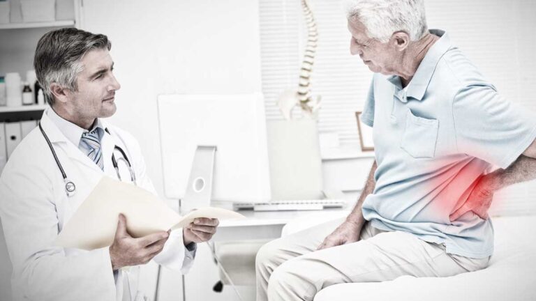 Ein alter Mann mit Rückenschmerzen im Gespräch mit einem Arzt in seiner Ordination. (c) AdobeStock