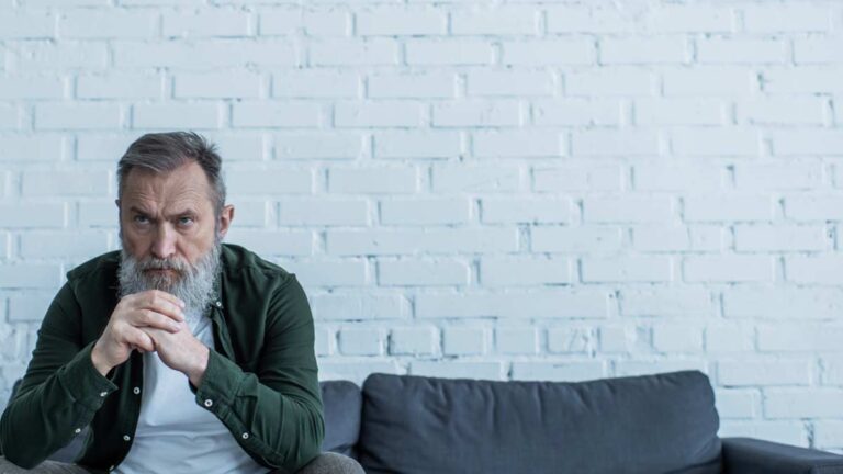 Ein alter Mann mit Bart sitzt traurig auf einer Couch. (c) AdobeStock
