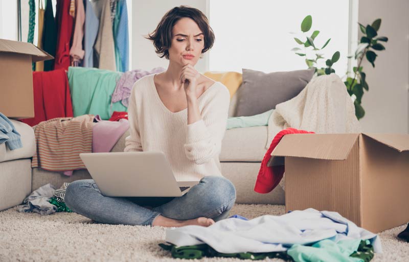 7 einfache Tipps für weniger Abfall und Mist beim Online-Kleiderkauf 1 Eine Frau mit Laptop sitzt am Boden neben unzähligen Kleidungsstücken und überlegt, Stichwort Kleiderkauf.
(c) AdobeStock