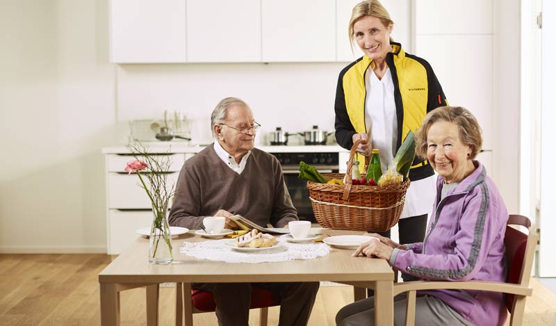 Ein älteres Paar am Tisch in einer Küche; eine Pflegerin bringt mit einem Korb Essen.
(c) Hilfswerk Oesterreich/ Johannes Puch