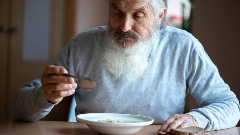 Ein älterer Mann mit weißem Vollbart an einem Tisch beim Suppe Essen. (c) AdobeStock