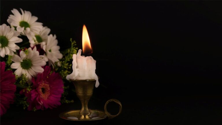 Eine brennende Kerze neben einem Blumenstrauß. (c) AdobeStock