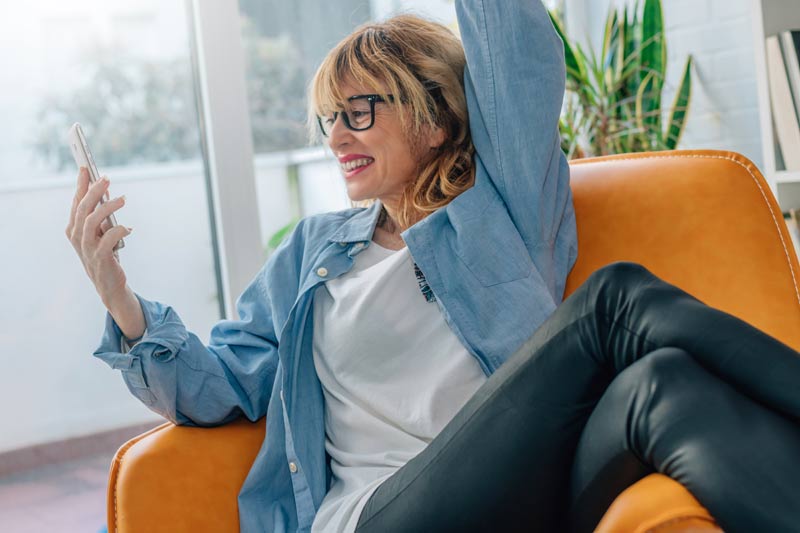 Eine Frau mit Brille sitzt in einem großen Ledersessel, hält ein Smartphone in der Hand und jubelt/ freut sich.
(c) AdobeStock