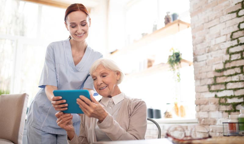 Eine alter Frau mit Tablett, neben ihr eine Pflegerin, die ihr das Gerät erklärt, Stichwort Wohnen im Alter.
(c) AdobeStock