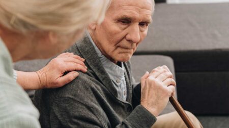 Ein alter Mann mit Demenz auf einer Couch. Eine Frau legt ihm die Hand auf die Schulter. (c) AdobeStock