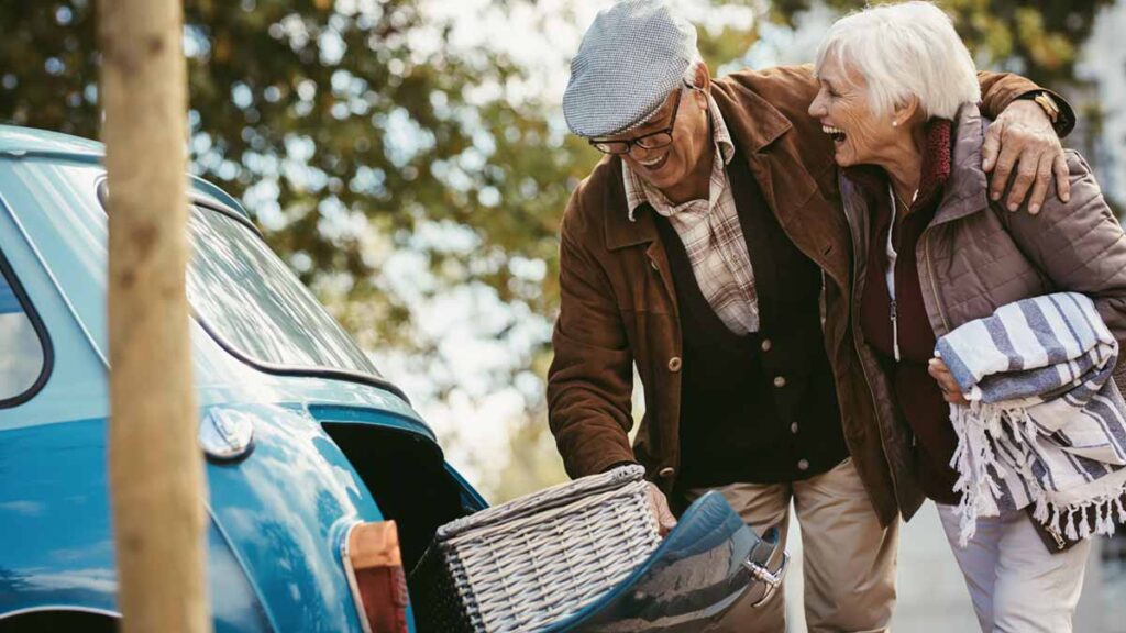 Ein älteres Paar mit einem Picknickkorb, den der Mann aus einem Auto holt. (c) AdobeStock