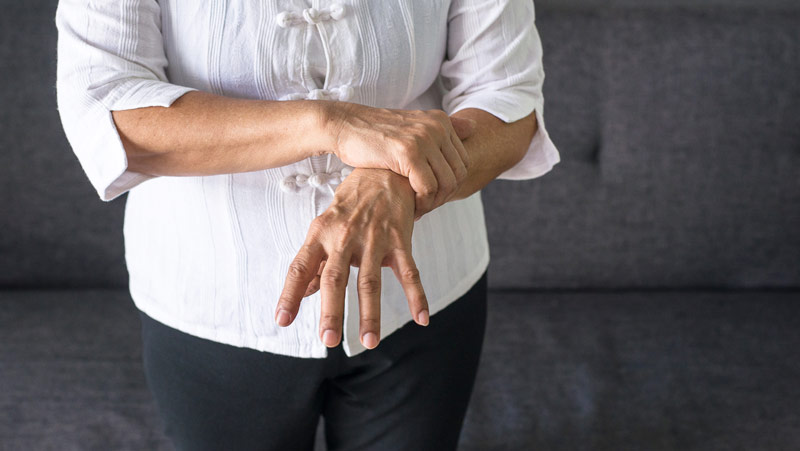 Eine alte Frau, die mit einer Hand den Unterarm ihrer anderen Hand hält, Stichwort Morbus Parkinson.
(c) AdobeStock