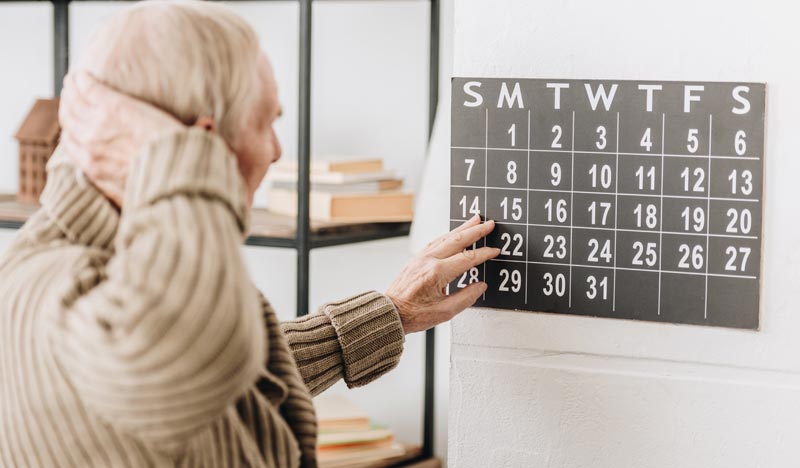 Ein alter Mann, der verwirrt vor einem Kalender steht, eine Hand am Kalender, die andere an seinem Hinterkopf, Stichwort Demenz-Prävention.
(c) AdobeStock