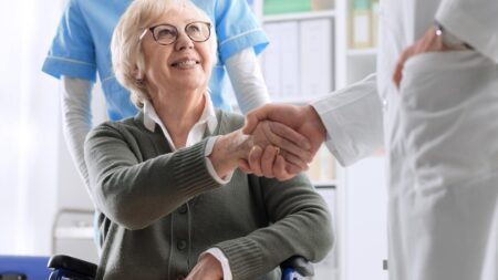 Eine ältere Frau in einem Rollstuhl gibt einem Arzt die Hand. (c) AdobeStock