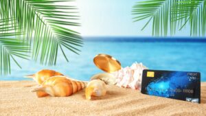 AK warnt: Spesenfalle „Urlaubsgeld“! Bis zu 20 Euro Spesen beim Abheben mit der Kreditkarte Eine Bankomatkarte neben Muscheln im Sand, dahinter Meer. (c) AdobeStock