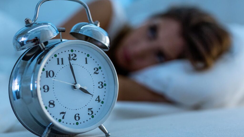 Ein Wecker im Vordergrund, dahinter eine Frau in einem Bett, die nicht schlafen kann. (c) AdobeStock