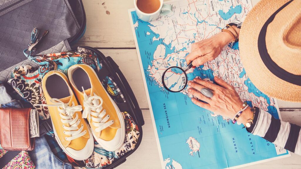 Die Hände einer älteren Frau mit Sonnenhut, die mit einer Lupe auf einer Landkarte schaut; daneben Reiseutensilien. (c) AdobeStock
