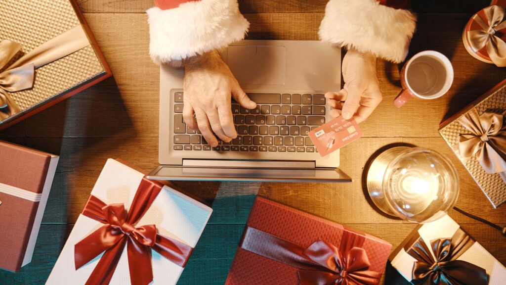 Von oben: ein Tisch, an dem eine Person im Weihnachtskostüm an einem Laptop sitzt und seine Kreditkartendetails eingibt; um den Laptop liegen Weihnachtsgeschenke am Tisch. (c) AdobeStock