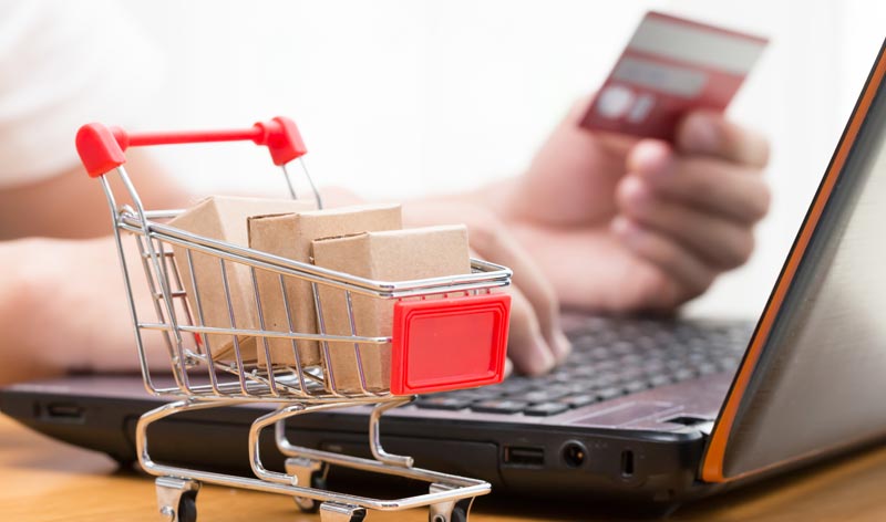 Die Hände einer Frau, die eine Kreditkarte hält und die Daten in einem Laptop eingibt; davor ein kleiner Einkaufswagen mit drei kleinen Päckchen, Stichwort Online-Shop.
(c) AdobeStock