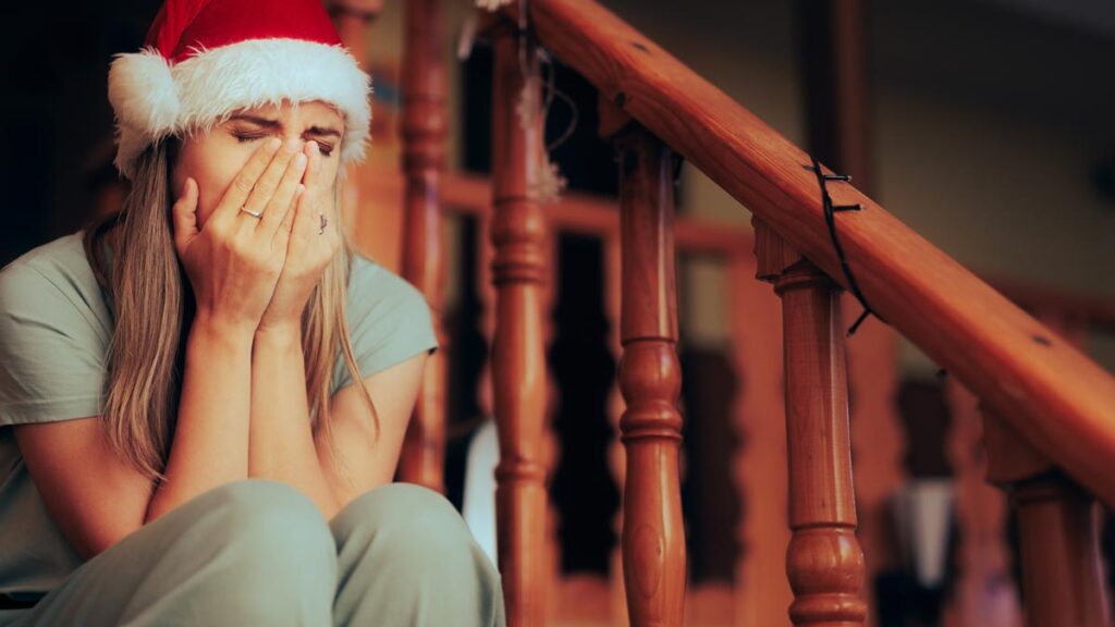 Eine Frau mit Weihnachtsmütze sitzt auf einer Treppe und hält sich die Hände vors Gesicht. (c) AdobeStock