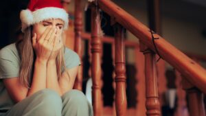 Eine Frau mit Weihnachtsmütze sitzt auf einer Treppe und hält sich die Hände vors Gesicht. (c) AdobeStock