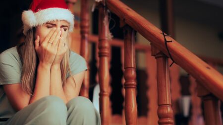 Eine Frau mit Weihnachtsmütze sitzt auf einer Treppe und hält sich die Hände vors Gesicht. (c) AdobeStock