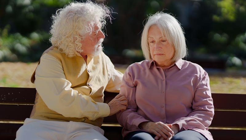 Ein älteres Paar auf einer Bank im Freien. Der Mann wendet sich zu der Frau, die apathisch ins  Leere schaut, Stichwort Demenz neu denken.(c) AdobeStock