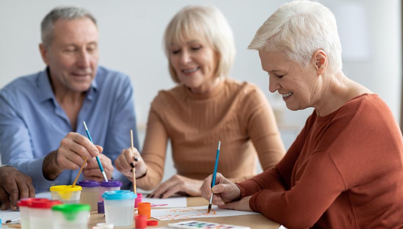 Drei Senioren malen gemeinsam mit Pinseln und Wasserfarben an einem Tisch und unterhalten sich lächelnd.(c) AdobeStock