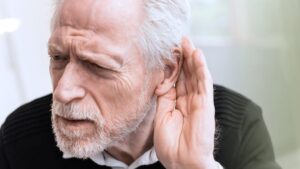 Älterer Mann mit grauem Bart hält sich die Hand ans Ohr und blickt angestrengt, um etwas besser zu verstehen – Symbol für beginnenden Hörverlust im Alter. (c) AdobeStock