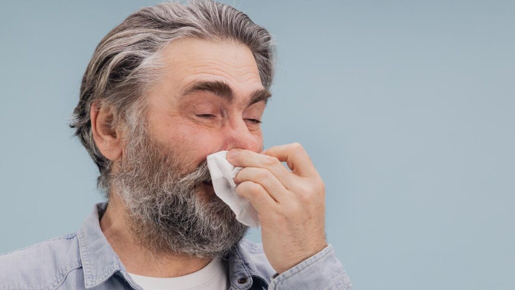 Pollensaison 2026: Warum die Frühblüher heuer intensiver zurückkehren Älterer Mann mit grauen Haaren und Bart nutzt ein Taschentuch aufgrund von allergischem Schnupfen während der Pollensaison. (c) AdobeStock