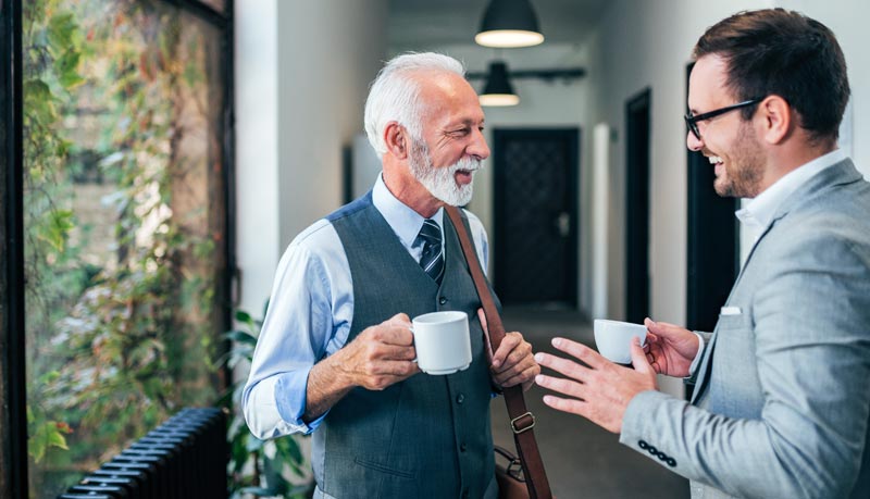 Ein älterer Mann und eine jüngerer mit Kaffeetassen in den Händen beim Plaudern auf dem Gang, Stichwort altersgemischte Teams.
(c) AdobeStock