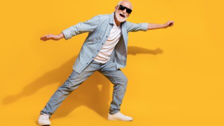 Ein modisch gekleideter älterer Mann mit weißem Bart, Sonnenbrille und Jeanshemd posiert dynamisch und lachend vor einem leuchtend gelben Hintergrund. (c) AdobeStock