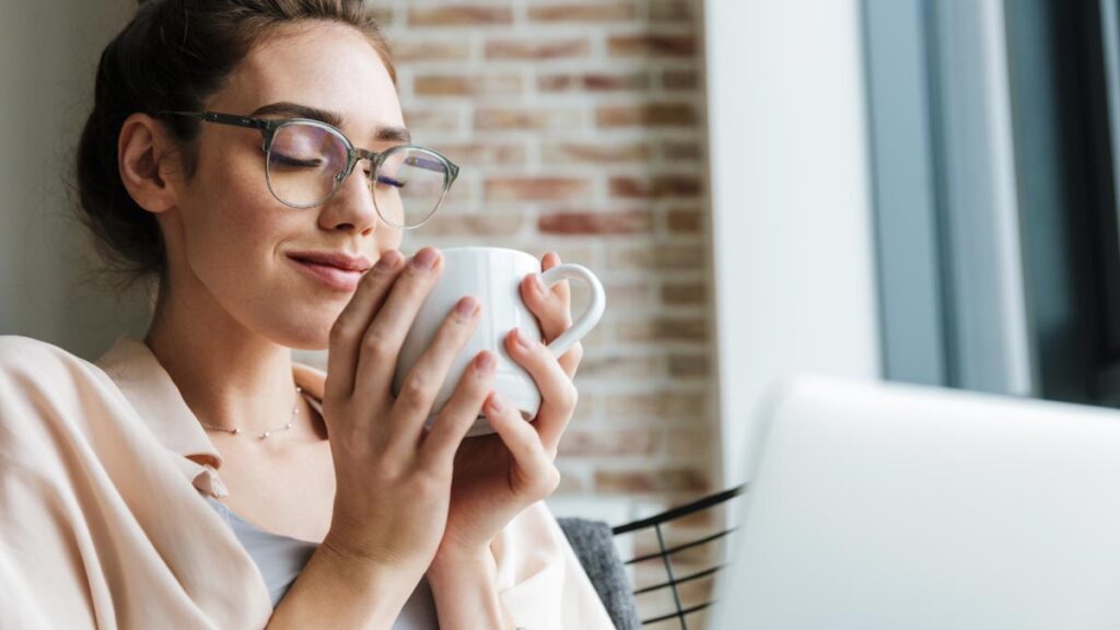 Brain-Food & Kaffee: Die 5 besten Kombinationen gegen Entzündungen Eine junge Frau mit Brille hält lächelnd und mit geschlossenen Augen eine weiße Kaffeetasse an ihr Gesicht, im Hintergrund eine Ziegelwand. (c) AdobeStock