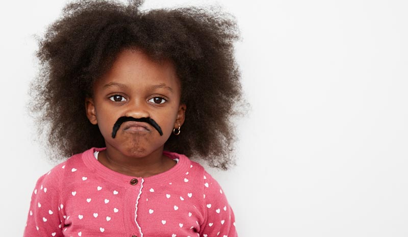 Ein kleines Mädchen mit voluminösem Haar und rosa Strickjacke schaut ernst in die Kamera, während sie einen aufgeklebten, schwarzen Schnurrbart trägt.(c) AdobeStock