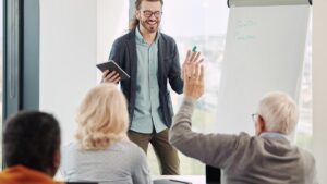 Ein lachender Dozent mit Tablet steht vor einer Gruppe von Senioren an einem Flipchart; ein Teilnehmer hebt meldend die Hand. (c) AdobeStock