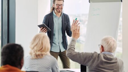Lebenslanges Lernen: Warum unser Gehirn keine Rente kennt Ein lachender Dozent mit Tablet steht vor einer Gruppe von Senioren an einem Flipchart; ein Teilnehmer hebt meldend die Hand. (c) AdobeStock
