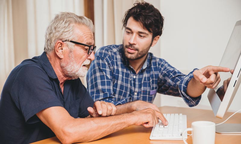 Ein jüngerer Mann erklärt einem älteren Herrn mit Brille am Computerbildschirm eine Anwendung, Stichwort lebenslanges Lernen.(c) AdobeStock