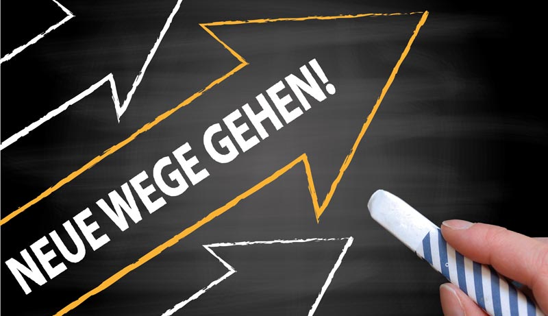 Eine Hand schreibt mit Kreide „NEUE WEGE GEHEN!“ auf eine Schiefertafel, auf der Pfeile nach oben zeigen.(c) AdobeStock