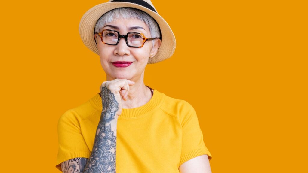 Aktives Altern durch bewusstes Reisen – Freizeit neu definiert Porträt einer stilvollen älteren Frau mit kurzem grauem Haar, Brille und Strohhut vor gelbem Hintergrund. Sie trägt ein gelbes Shirt, hat großflächige Tattoos am Arm und blickt selbstbewusst lächelnd in die Kamera. (c) AdobeStock