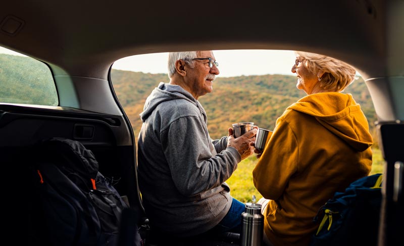 Aktives Altern durch bewusstes Reisen – Freizeit neu definiert 1 Ein älteres Paar sitzt entspannt im offenen Kofferraum eines Autos und blickt auf eine herbstliche Hügellandschaft. Sie halten Thermobecher in den Händen und unterhalten sich lächelnd; Wanderrucksäcke stehen bereit, Stichwort aktives Altern.(c) AdobeStock