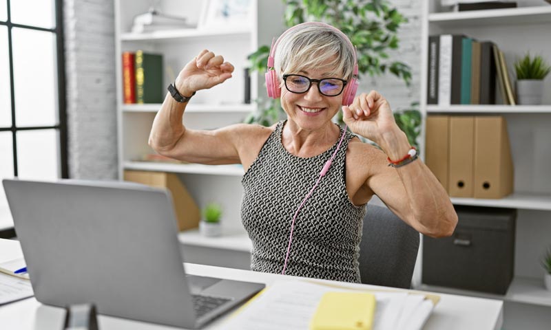 Gesundes Altern: Wie wir durch Lebenssinn und Teilhabe Vitalität gewinnen 2 Eine moderne ältere Frau mit Brille und rosa Kopfhörern sitzt lächelnd vor einem Laptop und hebt triumphierend die Arme.(c) AdobeStock