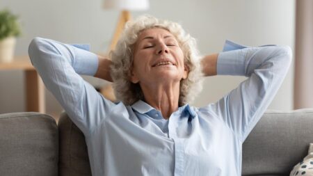 Eine ältere Frau mit grauen Locken lehnt sich entspannt auf einem Sofa zurück, die Hände hinter dem Kopf verschränkt und die Augen geschlossen – Sinnbild für mentale Gesundheit und Wohlbefinden. (c) AdobeStock