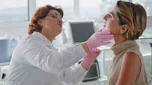 Eine Dermatologin in weißem Kittel und rosa Handschuhen untersucht vorsichtig das Gesicht und den Hals einer Patientin in einer hellen Praxis. (c) AdobeStock