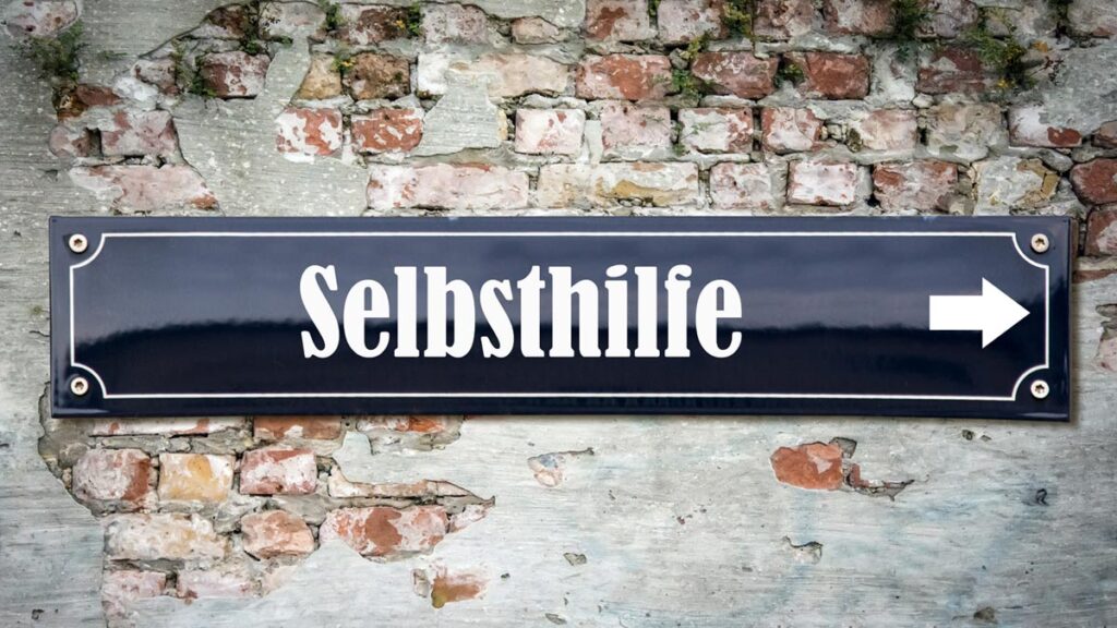 Dunkelblaues Straßenschild mit der weißen Aufschrift „Selbsthilfe“ und einem nach rechts weisenden Pfeil vor einer rustikalen Ziegelwand. (c) AdobeStock