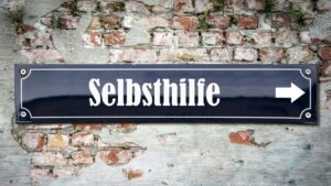 Dunkelblaues Straßenschild mit der weißen Aufschrift „Selbsthilfe“ und einem nach rechts weisenden Pfeil vor einer rustikalen Ziegelwand. (c) AdobeStock