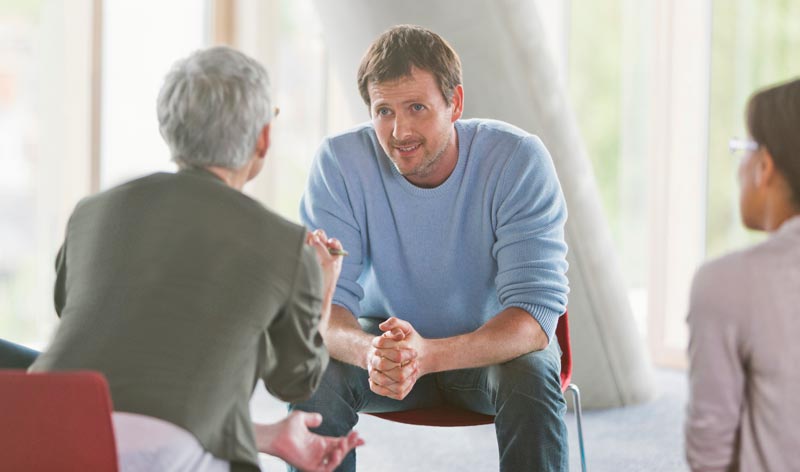 Ein Mann in einem blauen Pullover sitzt in einer Gesprächsrunde und hört einer Person mit grauen Haaren aufmerksam zu, Stichwort Selbsthilfegruppen Verzeichnis 2026.(c) AdobeStock