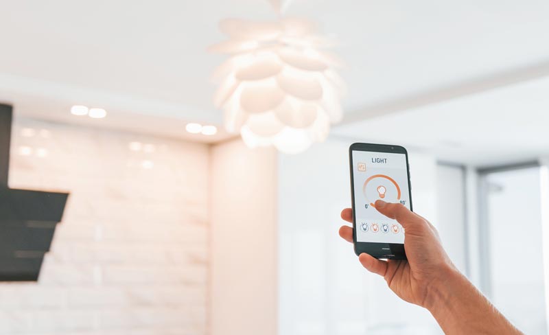 Smart Home für Senioren: 4 unschlagbare Vorteile für Ihre neue Freiheit 2 ine Person steuert über eine App auf dem Smartphone die Helligkeit einer modernen Designer-Deckenleuchte, Stichwort Smart Home für Senioren.(c) AdobeStock