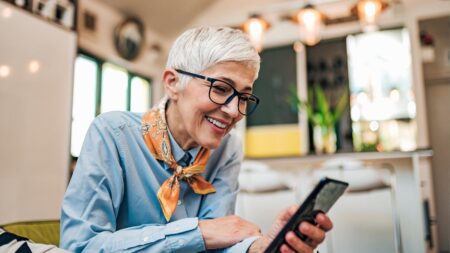 Eine lächelnde Seniorin mit Brille bedient kompetent ihr Smartphone in einer modernen, hellen Umgebung. (c) AdobeStock