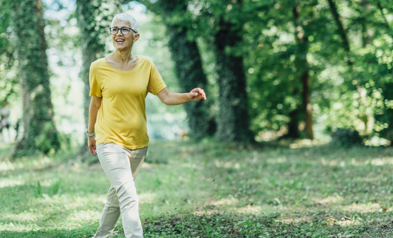 Eine aktive Frau mit Brille spaziert lächelnd durch einen sonnigen Wald und genießt ihre Lebensqualität, Stichwort Wechseljahre & Augen.(c) AdobeStock