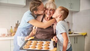 Eine lächelnde ältere Frau in einer Schürze hält ein Blech mit Keksen, während sie von zwei Kindern liebevoll auf die Wangen geküsst wird. (c) AdobeStock