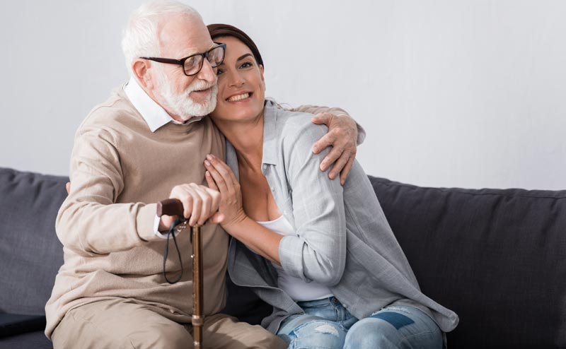 Eine lächelnde Frau umarmt einen älteren Mann mit Brille und Bart, der einen Gehstock hält, während beide auf einem Sofa sitzen.(c) AdobeStock