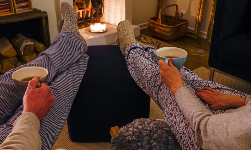 Zwei Personen sitzen entspannt mit Wollsocken und Teetassen vor einem brennenden Kamin – Inbegriff von Hygge und Geborgenheit.(c) AdobeStock