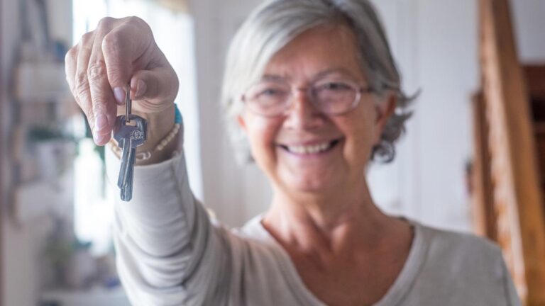 Eine lächelnde ältere Frau hält einen Hausschlüssel direkt in die Kamera, im Hintergrund ist ein heller Wohnraum mit Treppe zu erkennen. (c) AdobeStock