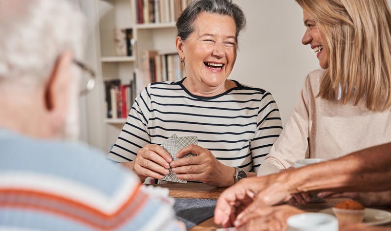 Eine Gruppe von Senioren sitzt lachend an einem Tisch und spielt Karten; im Fokus eine herzlich lachende Frau mit Kartenspiel in der Hand, Stichwort Wohnen im Alter.(c) AdobeStock