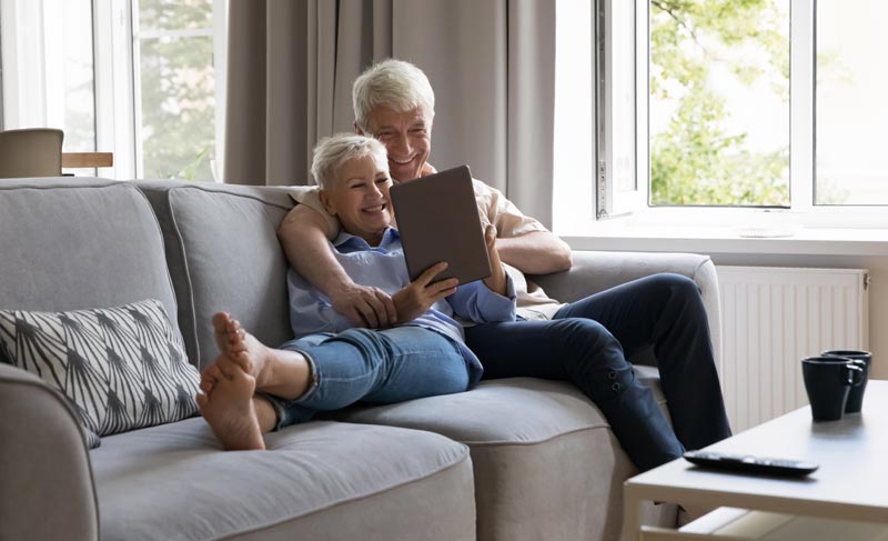 Ein älteres Paar sitzt entspannt auf einem grauen Sofa und blickt lächelnd auf ein Tablet, das sie gemeinsam in den Händen halten.(c) AdobeStock